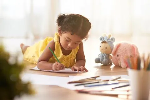 Young child in a yellow dress lying on the floor and coloring pages with colored pencils