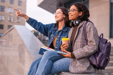 Two college students talking happily while sitting on a ledge pointing off to the distance